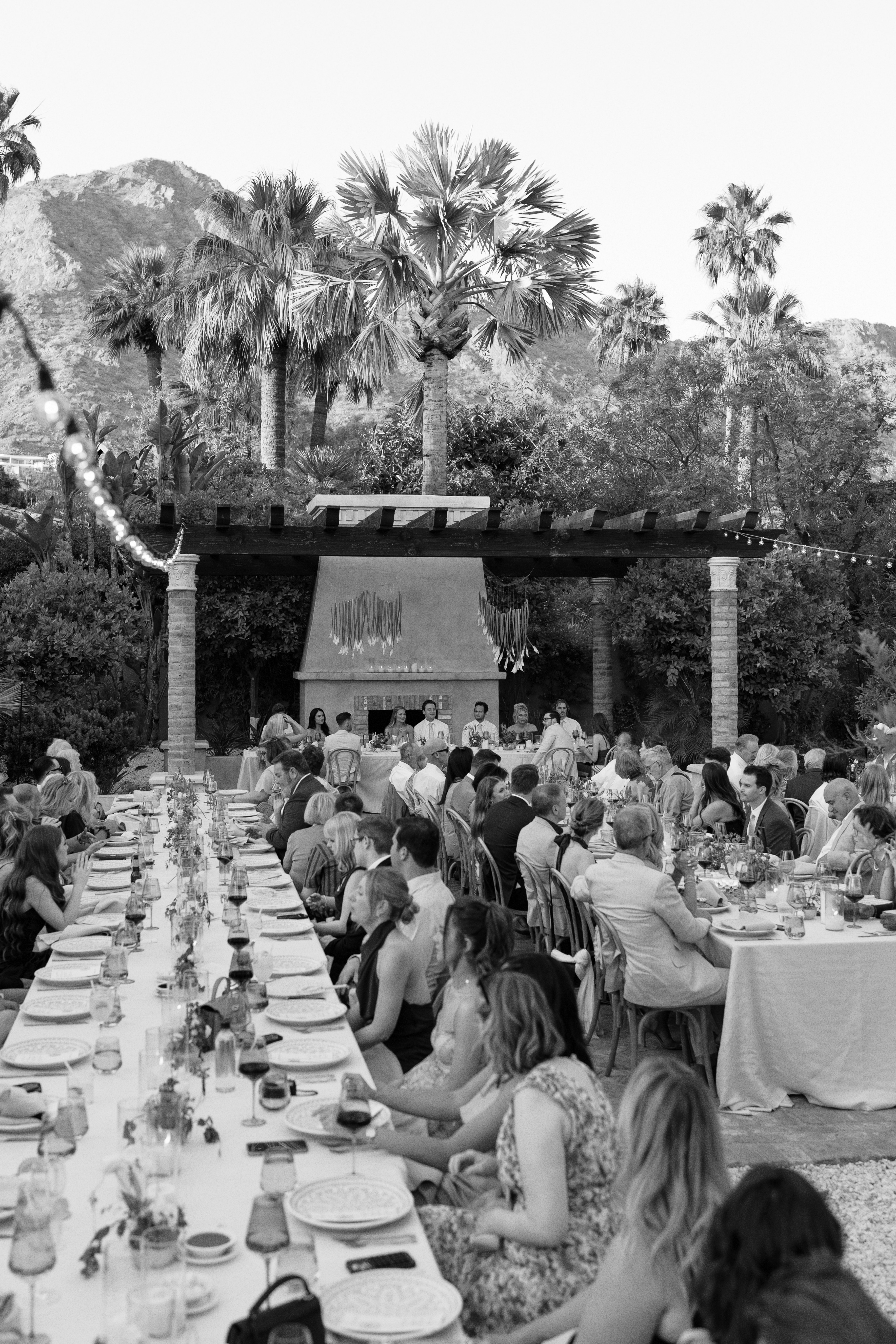 Outdoor desert wedding reception with guests seated at long banquet tables beneath a pergola, featuring palm trees and mountain views in the background.